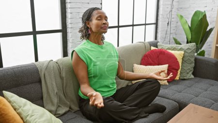Téléchargez les photos : Femme afro-américaine méditant paisiblement dans un salon moderne avec un canapé confortable et des plantes. - en image libre de droit