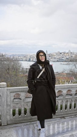 Téléchargez les photos : Une jeune femme dans un manteau élégant et des lunettes se tient devant un décor pittoresque istanbul, symbolisant le tourisme et la culture. - en image libre de droit