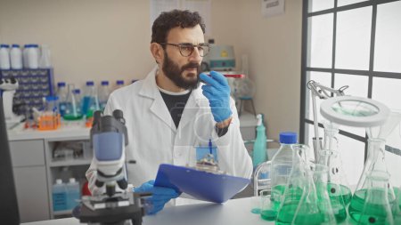 Téléchargez les photos : Homme barbu d'âge moyen en blouse de laboratoire examine un tube à essai à l'intérieur d'un laboratoire bien équipé avec presse-papiers à la main. - en image libre de droit
