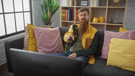 Foto de Un pelirrojo barbudo en ropa casual se sienta cómodamente en un sofá gris usando un control remoto dentro de una acogedora sala de estar con almohadas de colores. - Imagen libre de derechos