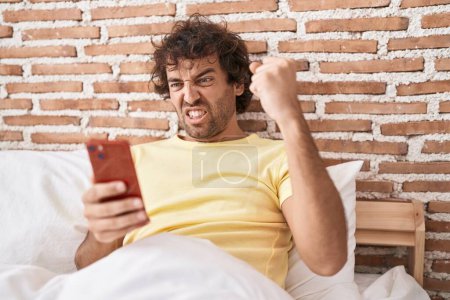 Photo for Hispanic young man using smartphone on the bed annoyed and frustrated shouting with anger, yelling crazy with anger and hand raised - Royalty Free Image