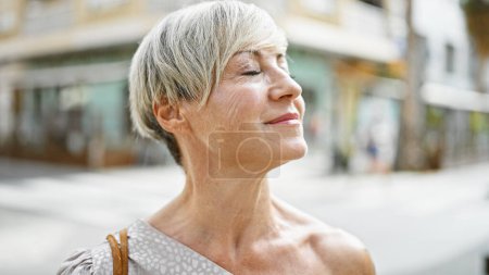 Photo for A mature woman with short grey hair enjoys sunlight on a busy urban street. - Royalty Free Image
