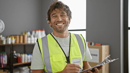 Téléchargez les photos : Homme souriant avec barbe portant gilet volontaire tenant presse-papiers dans l'entrepôt - en image libre de droit
