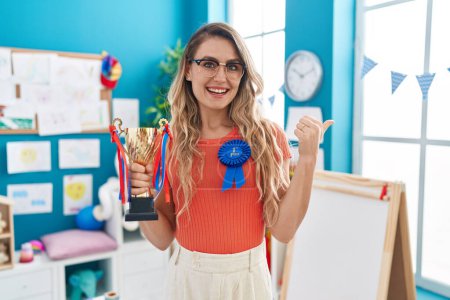 Photo for Young caucasian woman working as teacher at kindergarten holding prize pointing thumb up to the side smiling happy with open mouth - Royalty Free Image