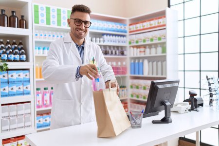 Téléchargez les photos : Jeune homme hispanique pharmacien mettre bouteille de médicaments sur le sac à provisions à la pharmacie - en image libre de droit