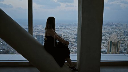 Téléchargez les photos : Une femme contemplative est assise à l'intérieur d'un observatoire surplombant un paysage urbain tentaculaire au crépuscule, incarnant la solitude et l'exploration urbaine.. - en image libre de droit