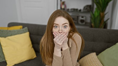 Foto de Una joven sorprendida con el pelo rubio cubriéndose la boca, sentada en un sofá en una acogedora sala de estar. - Imagen libre de derechos