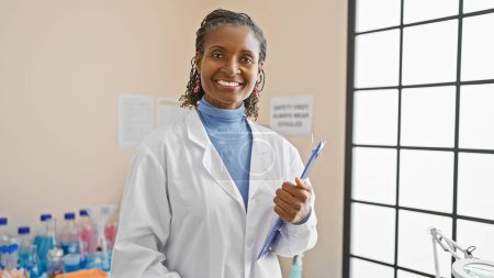 Téléchargez les photos : Femme scientifique afro-américaine confiante debout dans un laboratoire avec presse-papiers et stylo. - en image libre de droit