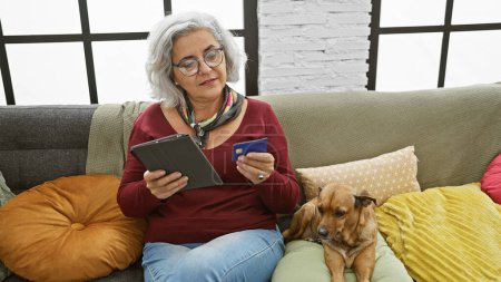 Téléchargez les photos : Femme mûre avec des lunettes à l'aide d'une tablette et tenant la carte de crédit, chien à côté d'elle, dans un salon confortable intérieur. - en image libre de droit