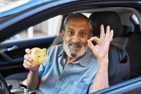 Téléchargez les photos : Homme âgé joyeux clignotant un signe ok confiant avec ses doigts, souriant d'oreille en oreille, comme il câline sa tirelire près d'une voiture fraîche dans les rues de la ville. un portrait du succès et du bonheur! - en image libre de droit