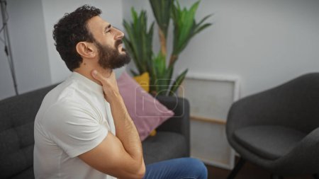 A bearded man in a white t-shirt experiencing neck pain in a modern living room