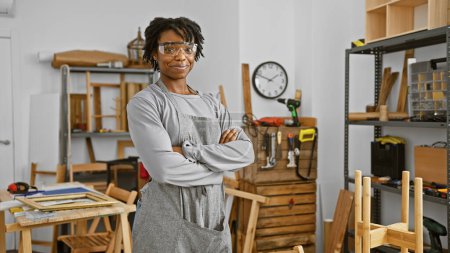 Téléchargez les photos : Femme confiante avec dreadlocks portant des lunettes de sécurité se tient dans un atelier de menuiserie. - en image libre de droit