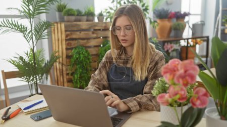 A young caucasian woman works on a laptop in a flower shop surrounded by plants and blooms.