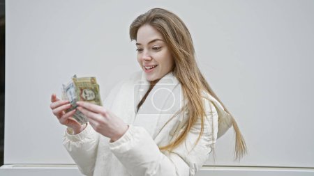 Foto de Mujer rubia sonriendo mientras examina sol moneda peruana sobre un fondo blanco, personifica las finanzas y la belleza. - Imagen libre de derechos