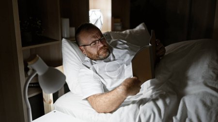 Attraktiver kaukasischer Mann mittleren Alters entspannt mit ernstem Gesichtsausdruck, verloren in der Welt eines guten Buches in seinem bequemen Bett, umgeben vom weichen Schein der Lampe in seinem gemütlichen Schlafzimmer