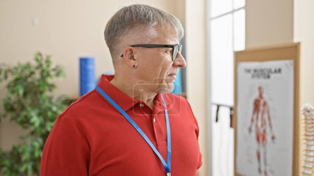 Photo for A mature grey-haired man in glasses wears a red shirt and lanyard beside a muscular system chart in a clinic. - Royalty Free Image