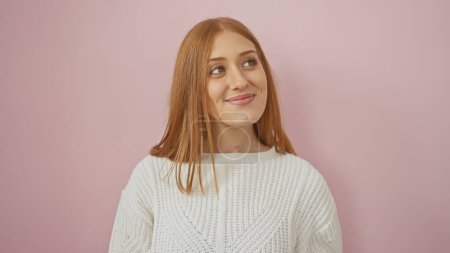 Young redhead woman in a white sweater poses with a subtle smile against a pink backdrop.