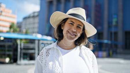 Foto de Hermosa joven hispana sonriendo confiada usando sombrero de verano en el edificio del Premio Nobel - Imagen libre de derechos