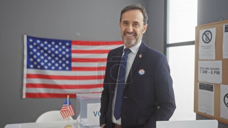 Mature man with beard stands proud in suit with 'i voted' sticker, american flag backdrop, at polling station.