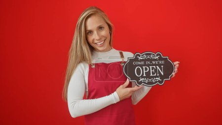 Téléchargez les photos : Une jeune femme souriante tenant un panneau "ouvert" sur fond rouge, représentant un service ou une entreprise accueillante. - en image libre de droit