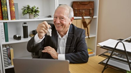 Photo for Smiling senior man, a successful real state agent, proudly holding keys of a new home in the office - Royalty Free Image