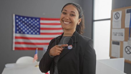 Téléchargez les photos : Une femme hispanique souriante pointe vers son autocollant "j'ai voté" dans un centre de vote universitaire avec un fond de drapeau américain - en image libre de droit