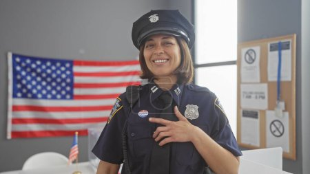 Téléchargez les photos : Une policière hispanique souriante avec un badge pointant vers son autocollant "j'ai voté" dans un centre de vote avec un drapeau américain. - en image libre de droit