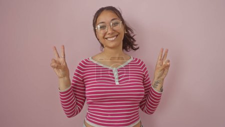 Foto de Una alegre joven hispana hace una señal de paz con ambas manos, de pie sobre un fondo rosa. - Imagen libre de derechos