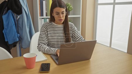 Téléchargez les photos : Jeune femme hispanique travaillant sur un ordinateur portable dans un cadre de bureau à domicile avec une tasse de café et smartphone sur le bureau. - en image libre de droit
