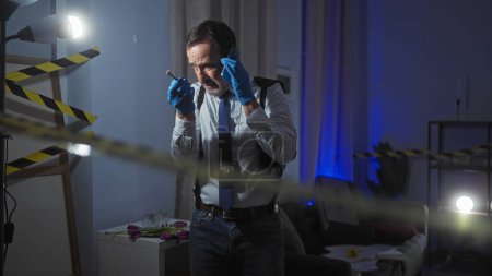 Mature man in suit holds phone and pen, analyzing clues in a dim, caution-taped indoor crime scene.