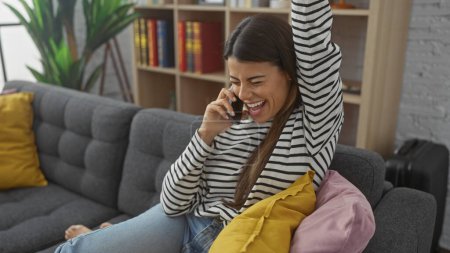 Téléchargez les photos : Une femme hispanique joyeuse bavardant sur un téléphone dans un salon confortable, respirant détente et bonheur. - en image libre de droit