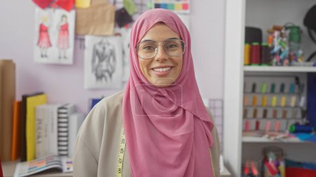 A smiling woman with a hijab and glasses in a colorful tailor shop with a measuring tape around her neck.