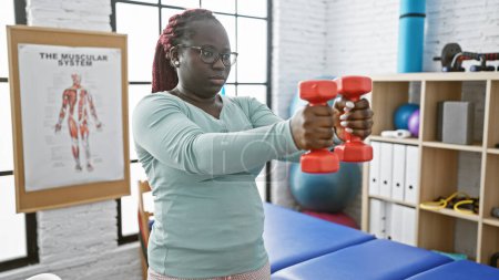 Photo for African american woman with braids lifting dumbbells in a physiotherapy clinic interior. - Royalty Free Image