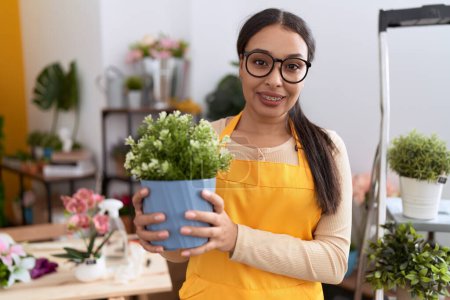 Téléchargez les photos : Jeune femme arabe travaillant au magasin de fleuriste tenant pot de plante regardant positif et heureux debout et souriant avec un sourire confiant montrant des dents - en image libre de droit