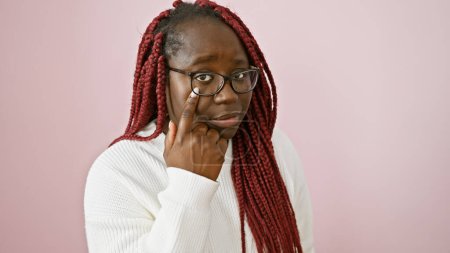 Thoughtful african woman with braids and glasses posing over a pink background