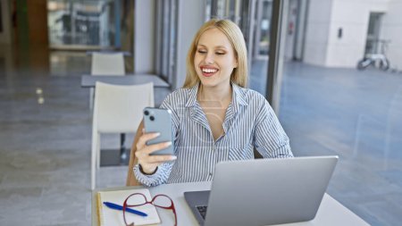Foto de Mujer rubia sonriente usando teléfono inteligente en una mesa de café con portátil y anteojos, entorno urbano en el fondo. - Imagen libre de derechos