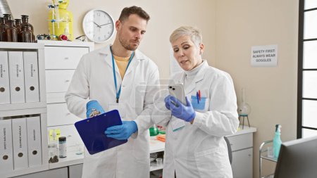 Photo for A man and woman in lab coats review data on a smartphone in a bright laboratory setting, portraying professionals at work. - Royalty Free Image