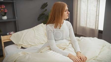 Foto de Una joven pensativa con el pelo rojo sentada en una cama en un dormitorio bien iluminado, mirando hacia otro lado pensativamente. - Imagen libre de derechos
