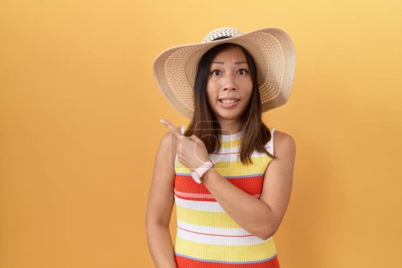 Foto de Mujer china de mediana edad con sombrero de verano sobre fondo amarillo señalando a un lado preocupado y nervioso con el dedo índice, preocupado y expresión sorprendida - Imagen libre de derechos