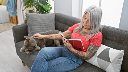 Entspannte Frau mittleren Alters, grauhaarige Bibliophile, kuschelt sich an ihren treuen Hund, sitzt gemütlich auf ihrem heimischen Sofa und genießt die Muße, in ihrem gemütlichen Wohnzimmer ein fesselndes Buch zu lesen..