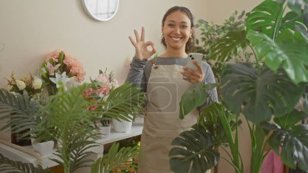 Téléchargez les photos : Femme hispanique souriante faisant signe correct dans la boutique de fleurs verte remplie de plantes tout en tenant smartphone - en image libre de droit