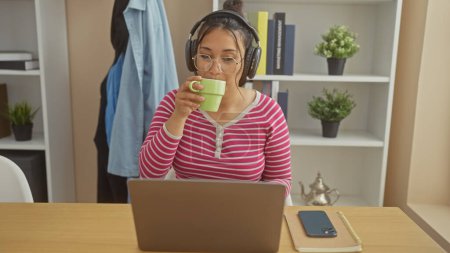 Hispanic woman drinking coffee while working on laptop at home, wearing headphones for concentration.