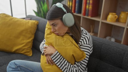 Photo for A pensive young woman wearing headphones cuddles a cushion on a sofa, exhibiting comfort and introspection in a cozy home setting. - Royalty Free Image