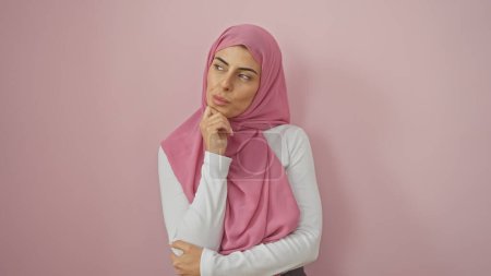 Foto de Una joven contemplativa en un hiyab rosado sobre un fondo rosa claro emana elegancia y curiosidad. - Imagen libre de derechos