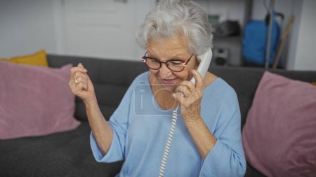 Photo for An elderly woman joyfully talking on a telephone while seated on a couch indoors. - Royalty Free Image