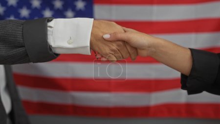 Foto de Un hombre y una mujer en atuendo formal estrechando manos en un ambiente interior con un fondo de bandera americana. - Imagen libre de derechos