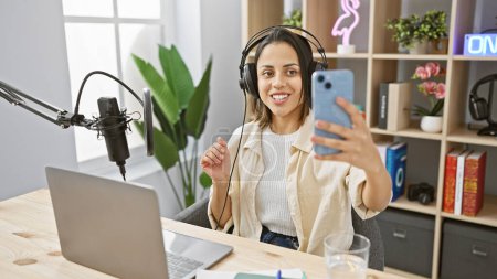 Una joven sonriente con auriculares se toma una selfie en una moderna configuración de estudio de radio, mostrando tecnología de medios.