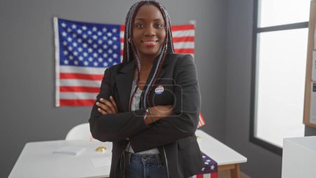 Mujer afroamericana con trenzas, brazos cruzados, vistiendo una pegatina de "yo voté", frente a una bandera americana en una universidad.