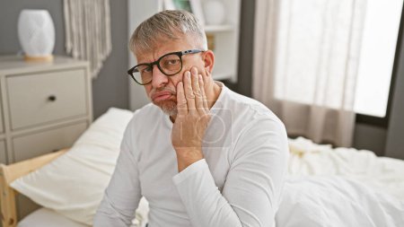 Photo for Middle-aged man with glasses seated on a white bed in a well-lit bedroom, appearing contemplative. - Royalty Free Image