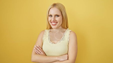Téléchargez les photos : Une jeune femme blonde souriante se tient les bras croisés sur un fond jaune, respirant confiance et beauté. - en image libre de droit